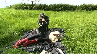 Fetish slave in latex walks and sucks a dildo on the grass